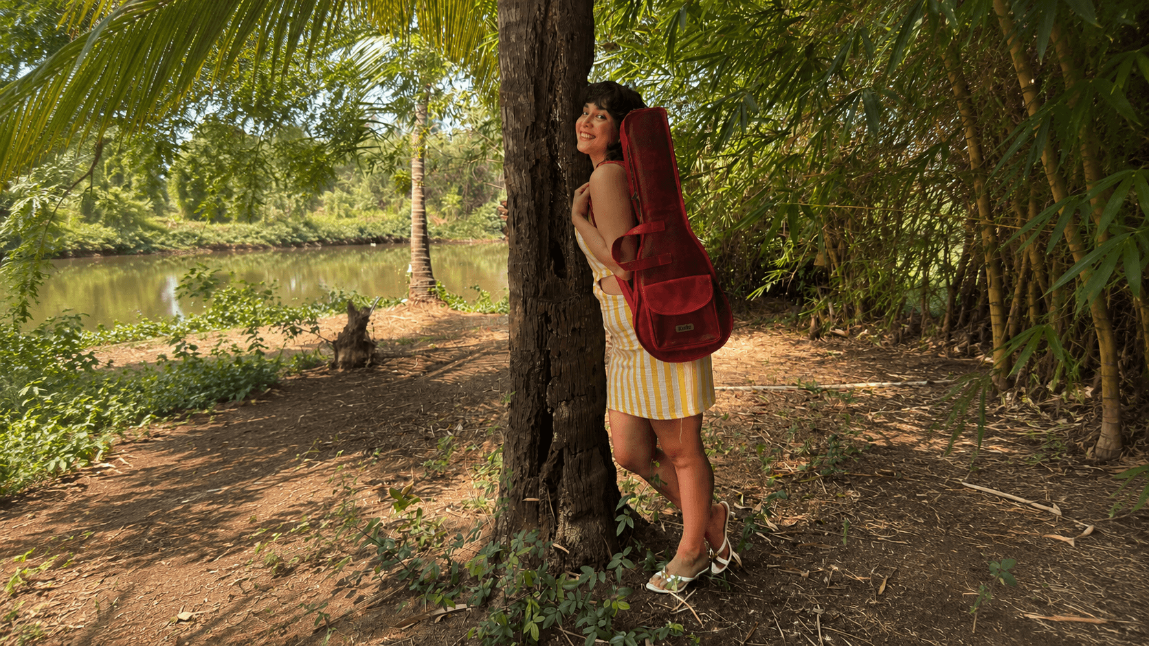 A woman by the river leaning on a tree with a red leather Kailo ukulele bag